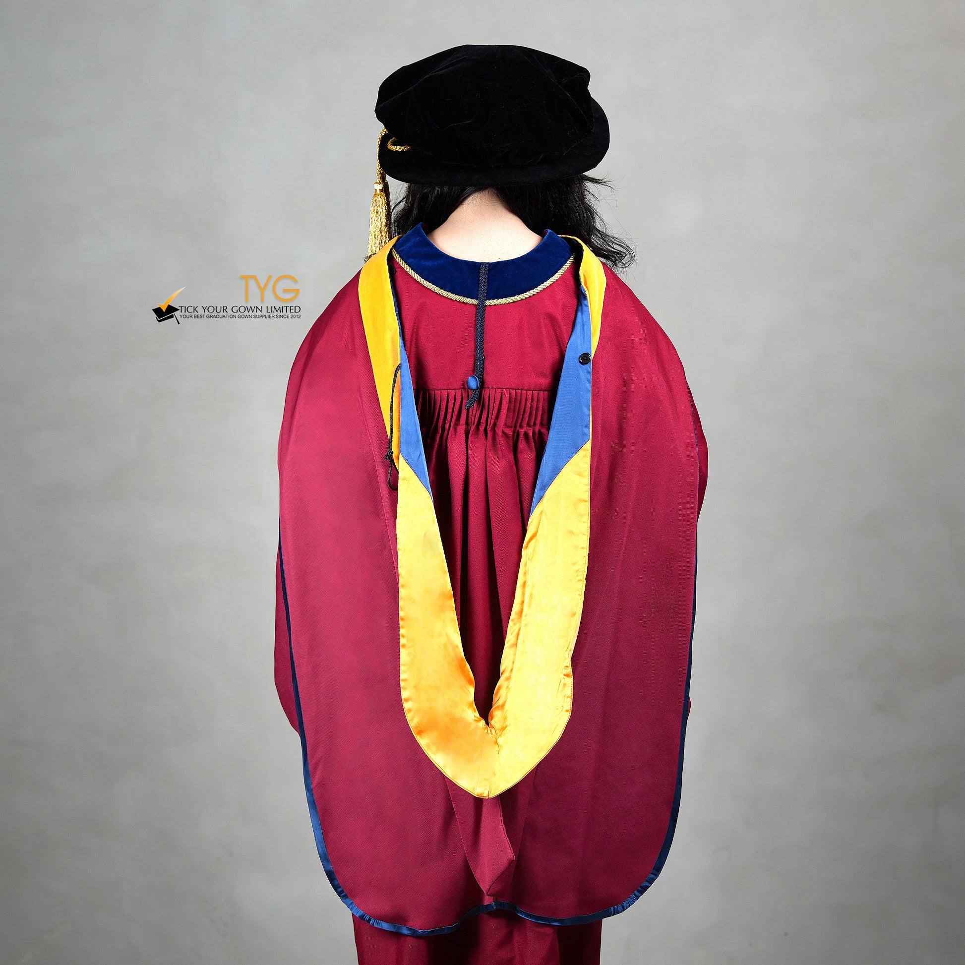 A back view of a person wearing a premium honorary graduation gown in maroon with royal blue velvet panels, gold trim, and a black doctoral bonnet with gold tassel