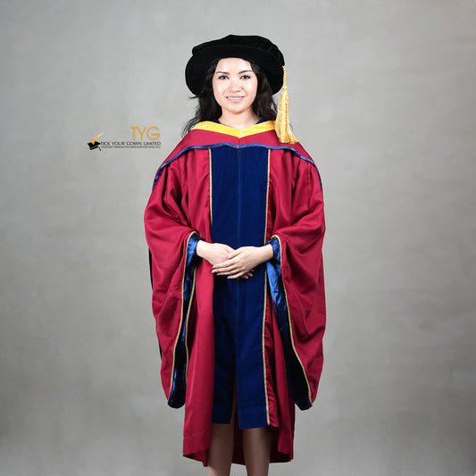 A front view of a person wearing a premium honorary graduation gown in maroon with royal blue velvet panels, gold trim, and a black doctoral bonnet with gold tassel