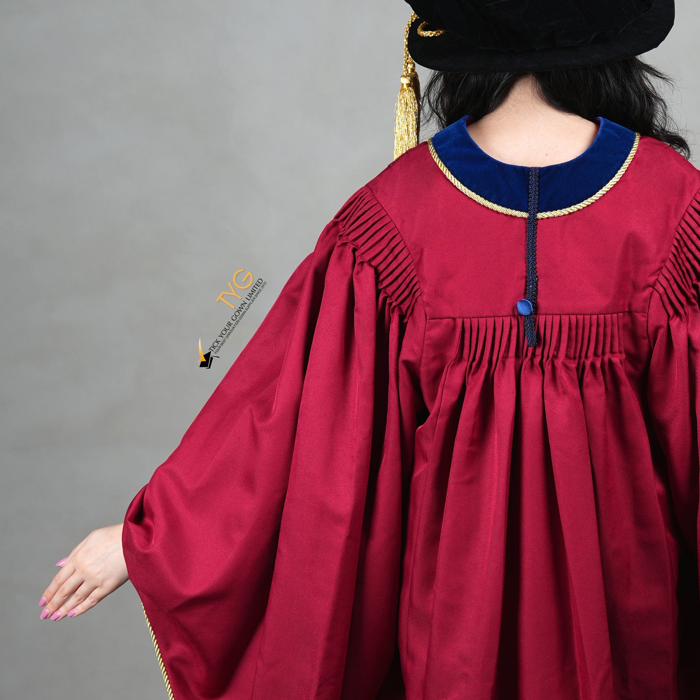 A detailed back view of a person wearing a premium honorary graduation gown in maroon with royal blue velvet panels, gold trim, and a matching doctoral bonnet with gold tassel.
