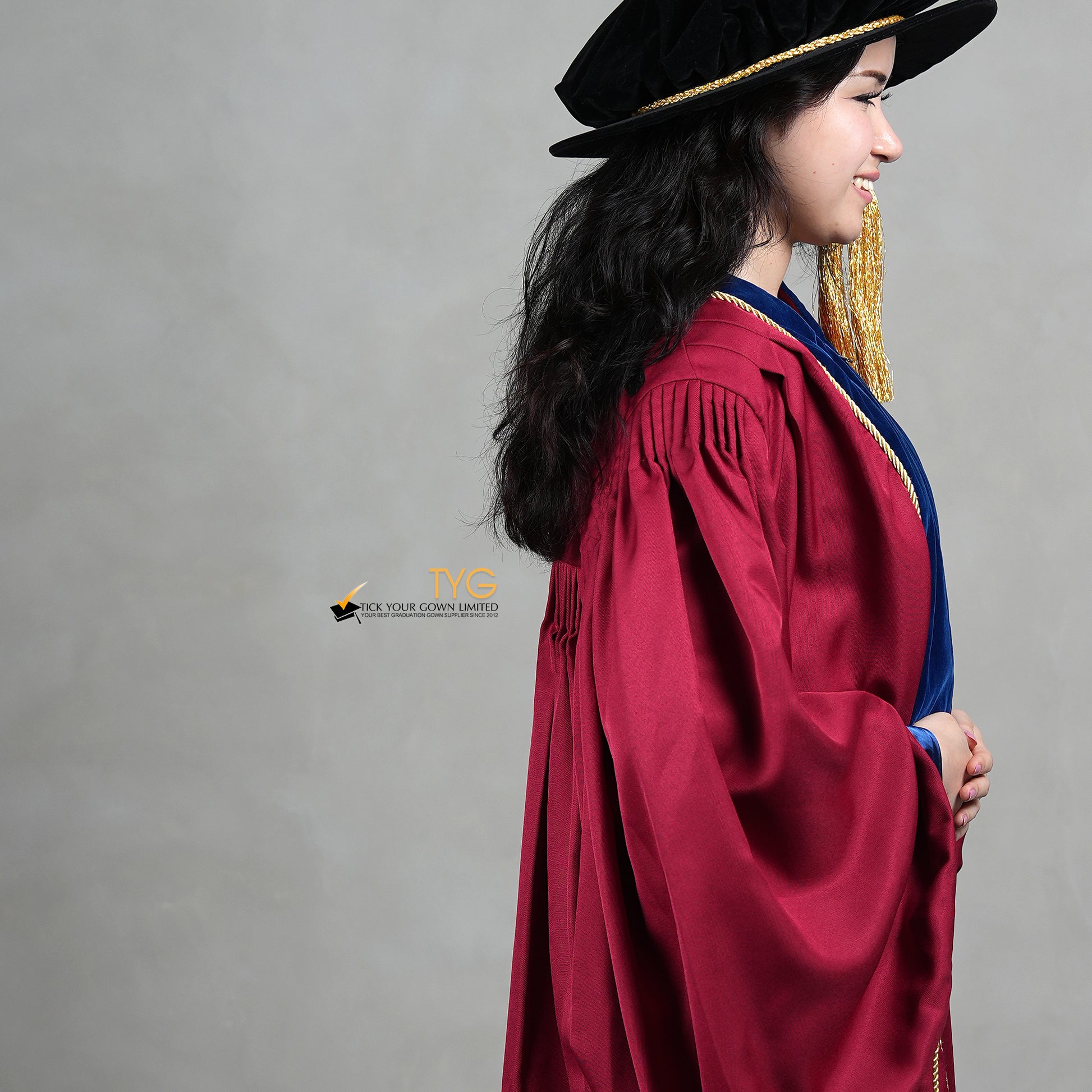 A detailed side view of a person wearing a premium honorary graduation gown in maroon with royal blue velvet panels, gold trim, and a matching doctoral bonnet with gold tassel.