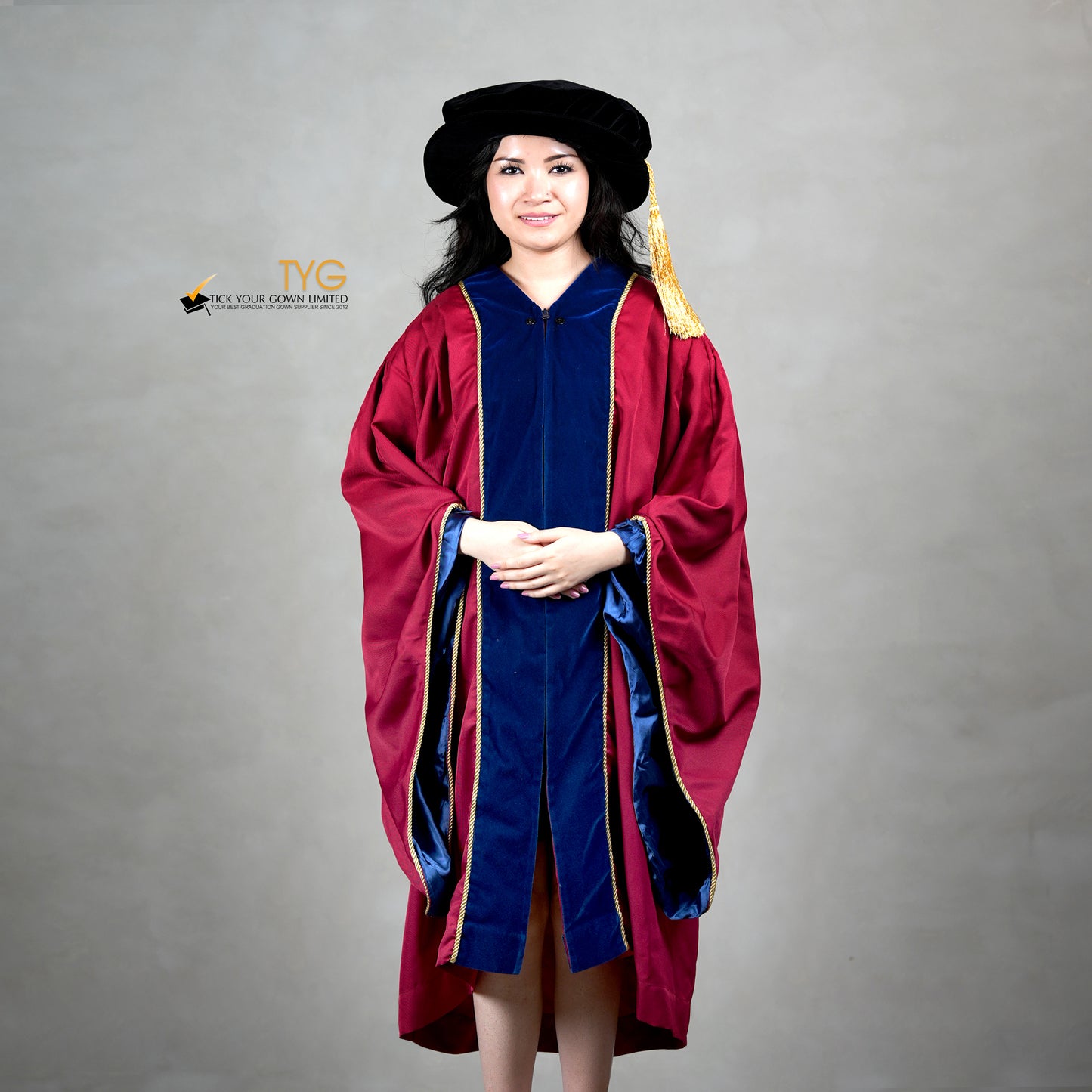 Front view of a person wearing a premium honorary graduation gown in maroon with royal blue velvet panels, gold trim, and a matching doctoral bonnet with gold tassel.