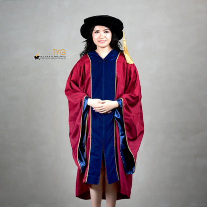 Front view of a person wearing a premium honorary graduation gown in maroon with royal blue velvet panels, gold trim, and a matching doctoral bonnet with gold tassel.