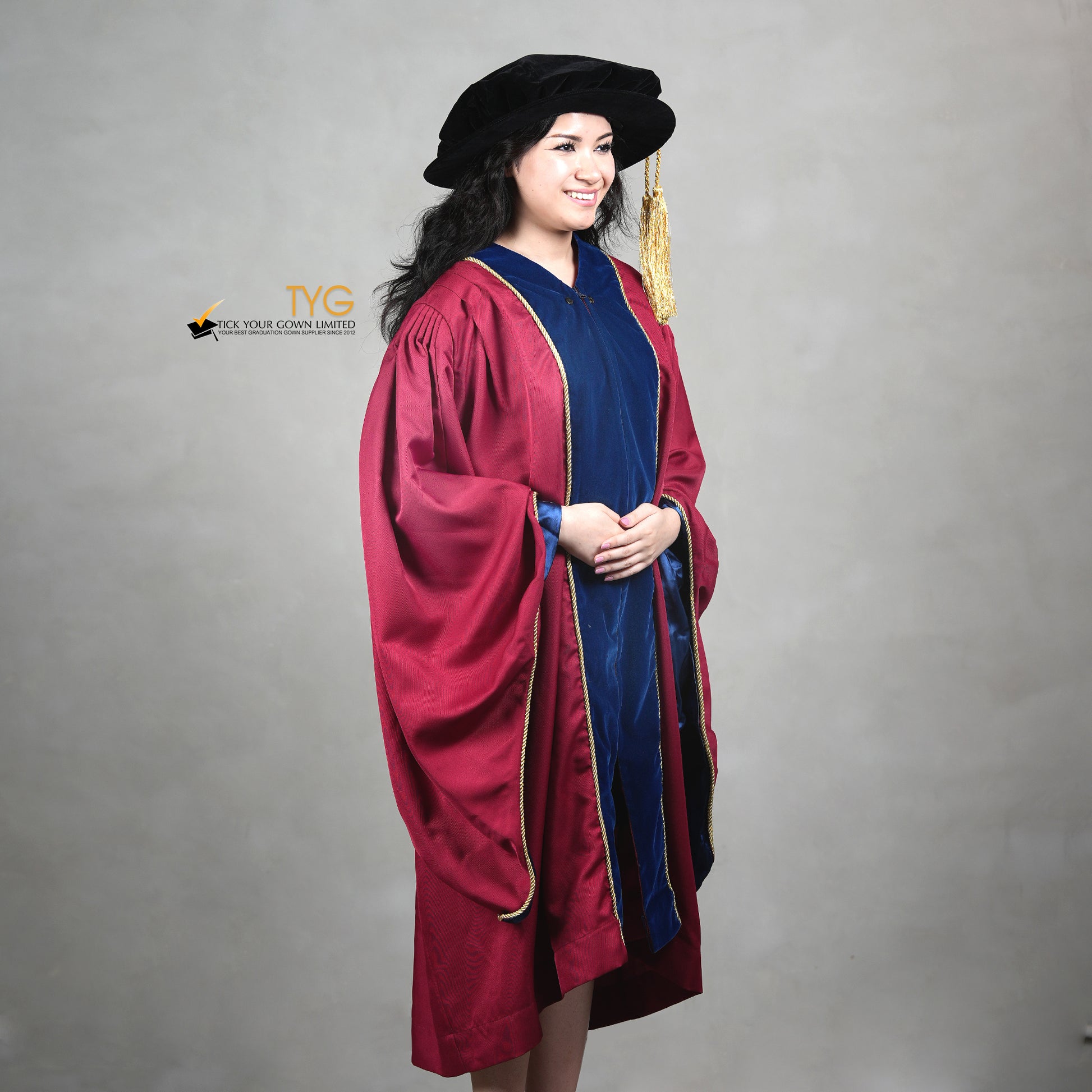 A profile view of a person wearing a premium honorary graduation gown in maroon with royal blue velvet panels, gold trim, and a matching doctoral bonnet with gold tassel.