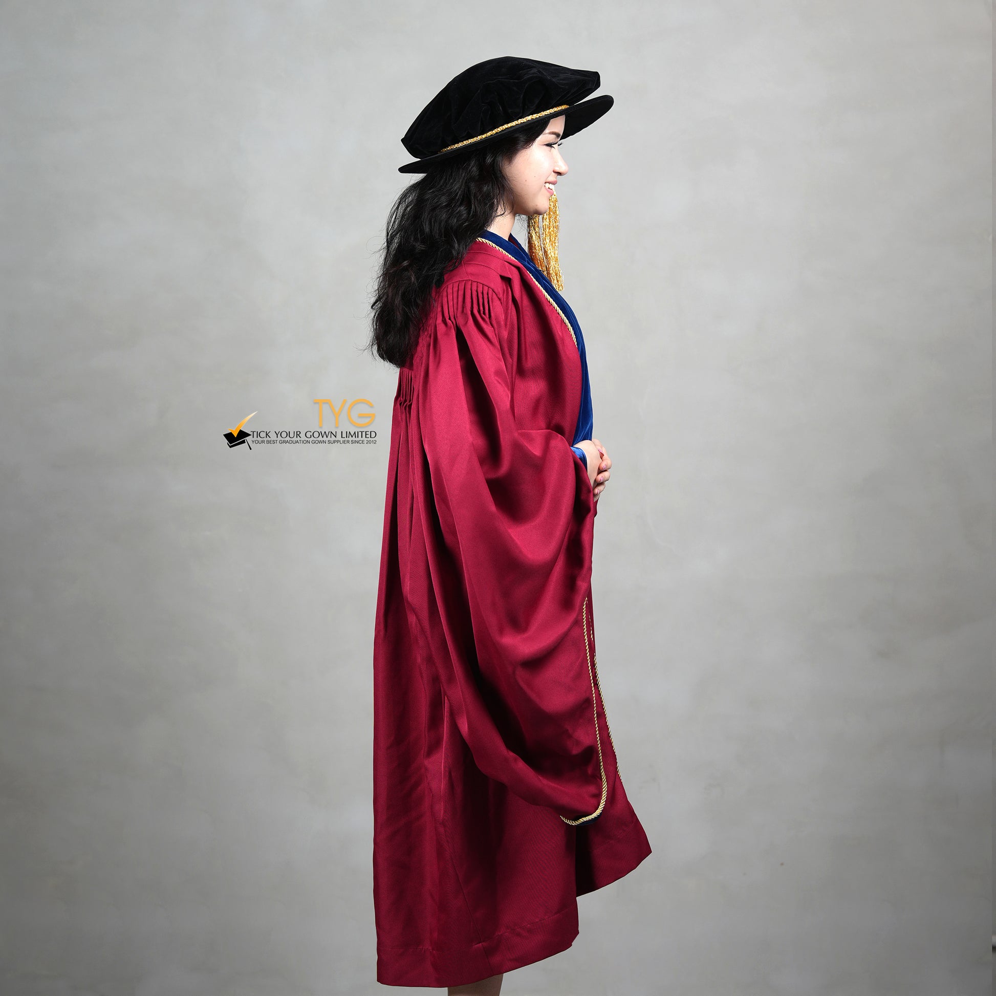 A side view of a person wearing a premium honorary graduation gown in maroon with royal blue velvet panels, gold trim, and a matching doctoral bonnet with gold tassel.