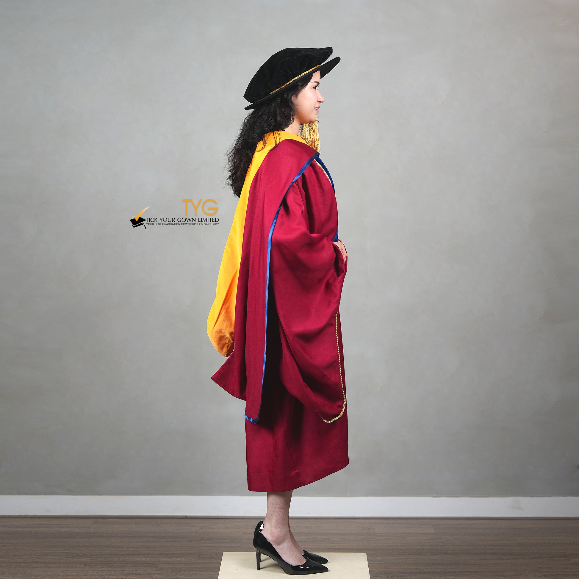 A side view of a person wearing a premium honorary graduation gown in maroon with royal blue velvet panels, gold trim, and a black doctoral bonnet with gold tassel
