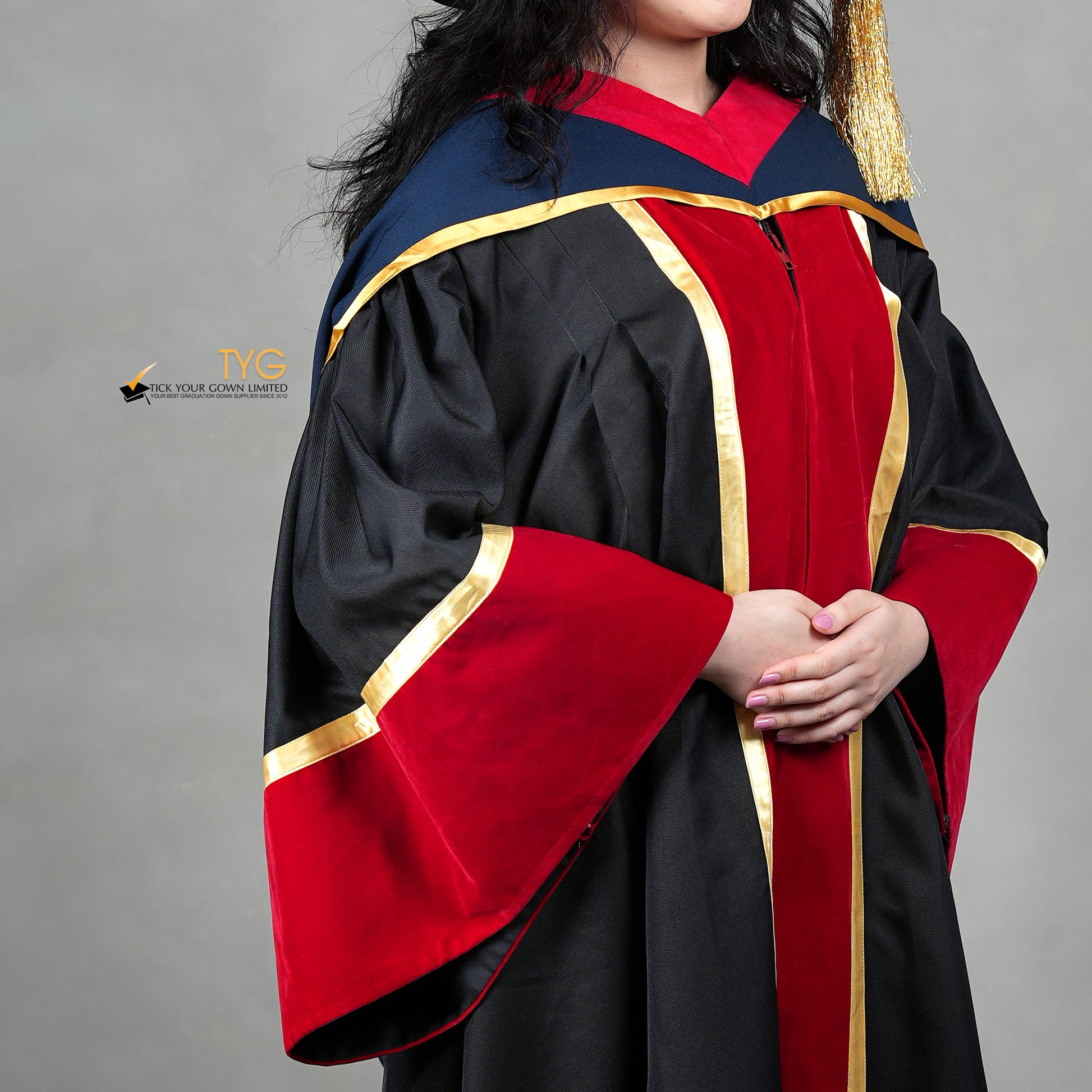 A detailed profile view of a person wearing a premium honorary graduation gown in black with red velvet panels, gold trim, matching red velvet sleeve cuffs, and a doctoral bonnet with gold tassel