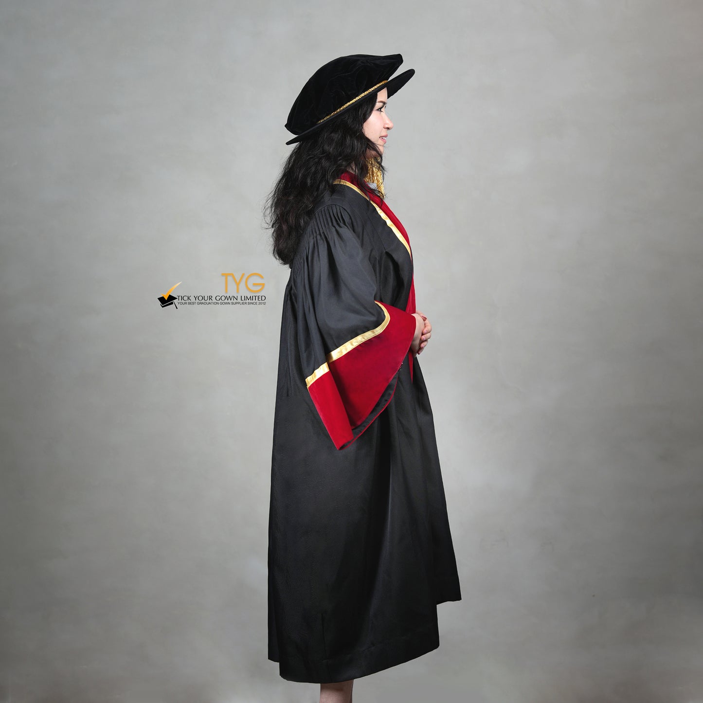 Profile view of a doctoral graduation gown in black with red velvet sleeve cuffs, gold trim, and a matching doctoral bonnet with gold tassel