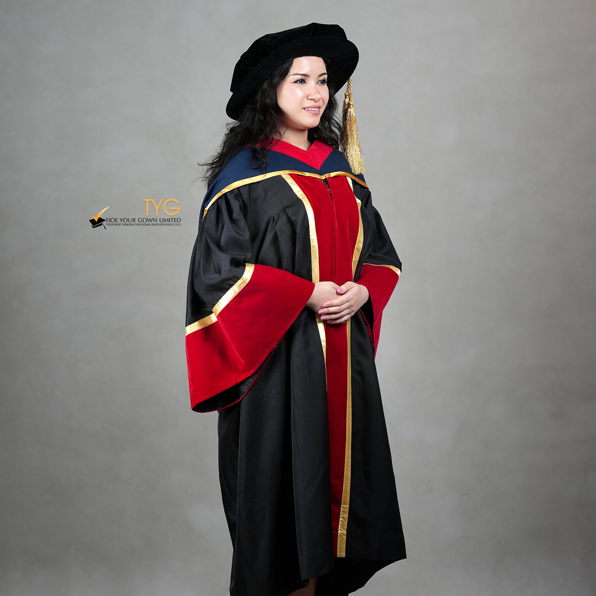 A profile view of a person wearing a premium honorary graduation gown in black with red velvet panels, gold trim, matching red velvet sleeve cuffs, and a doctoral bonnet with gold tassel