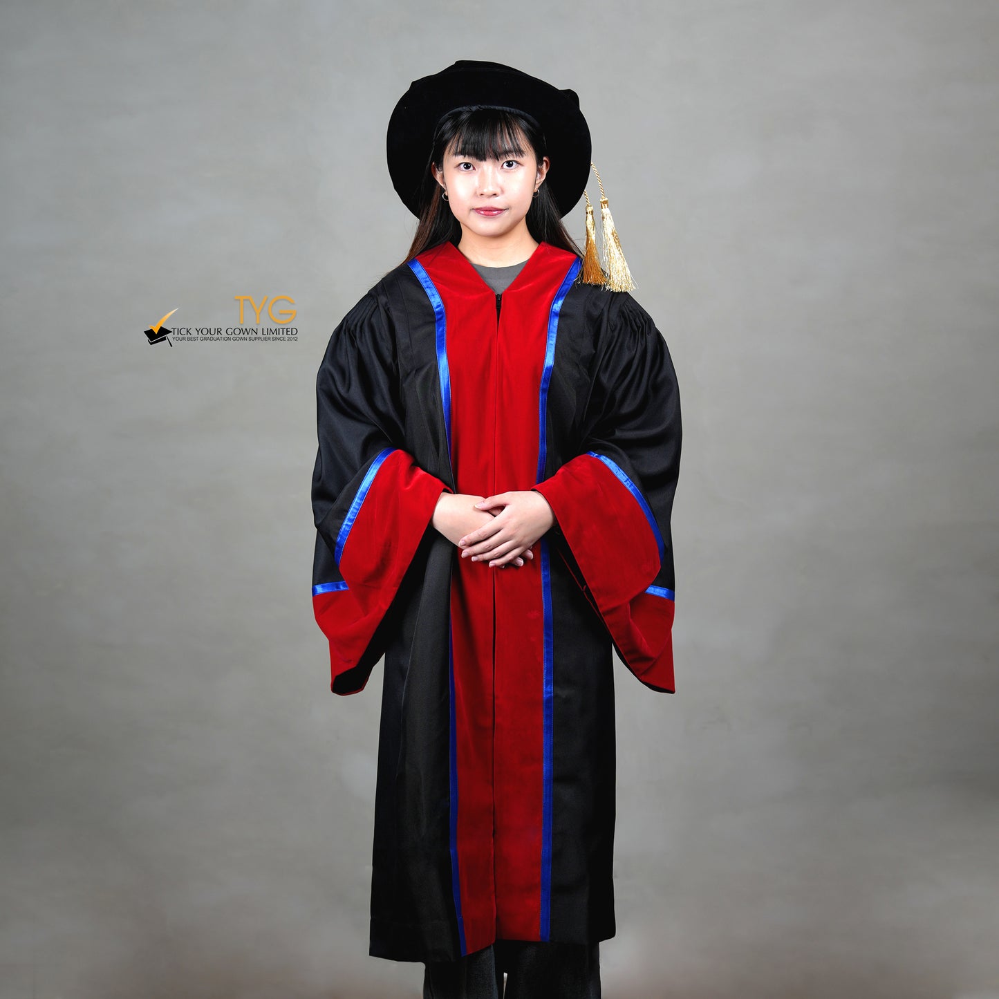 A front view of a person wearing a premium honorary graduation gown in black with red velvet panels, royal blue trim, red velvet sleeve cuffs, and a matching doctoral bonnet with gold tassel.