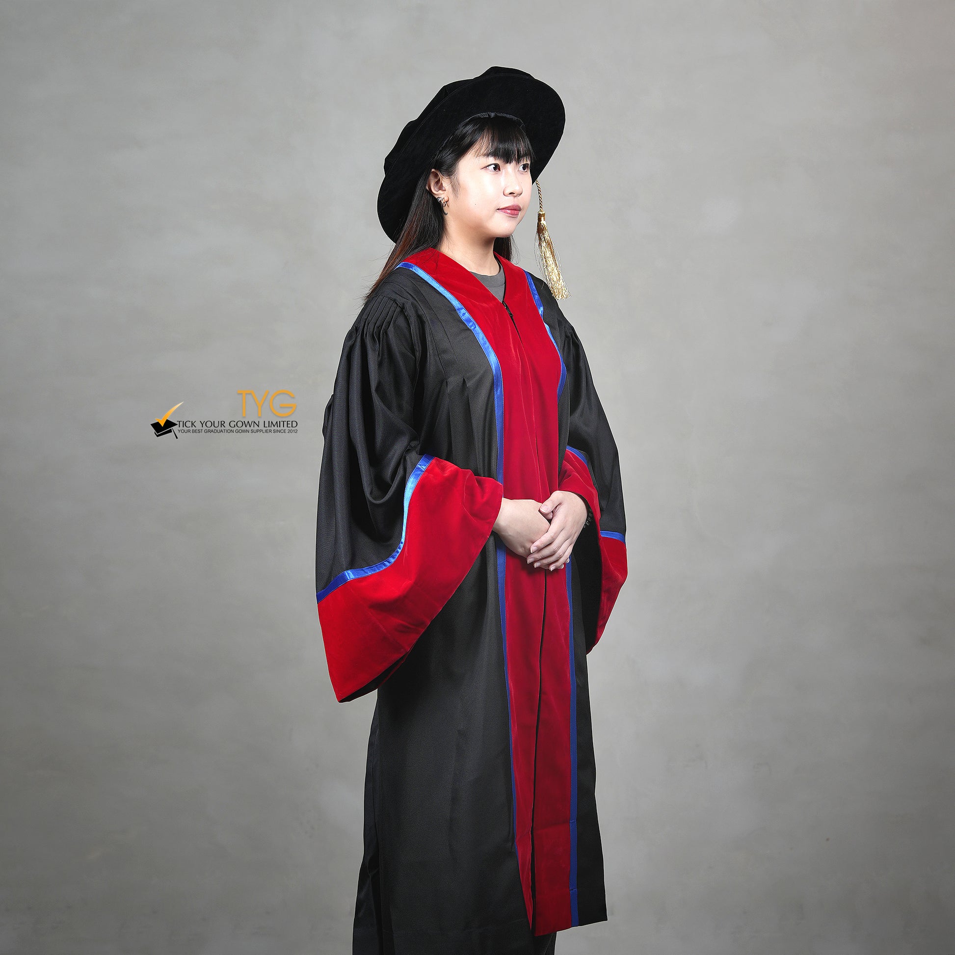 A profile view of a person wearing a premium honorary graduation gown in black with red velvet panels, royal blue trim, red velvet sleeve cuffs, and a matching doctoral bonnet with gold tassel.