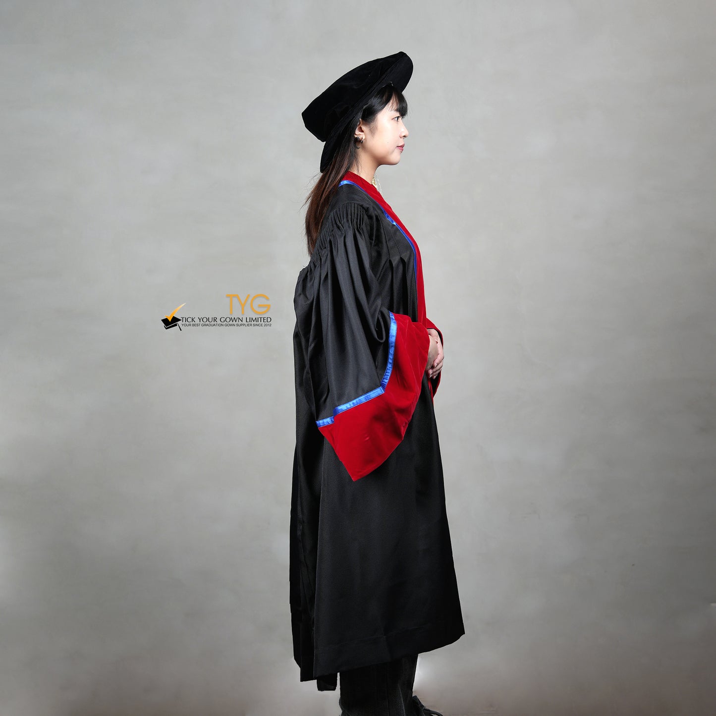 A side view of a person wearing a premium honorary graduation gown in black with red velvet panels, royal blue trim, red velvet sleeve cuffs, and a matching doctoral bonnet with gold tassel.