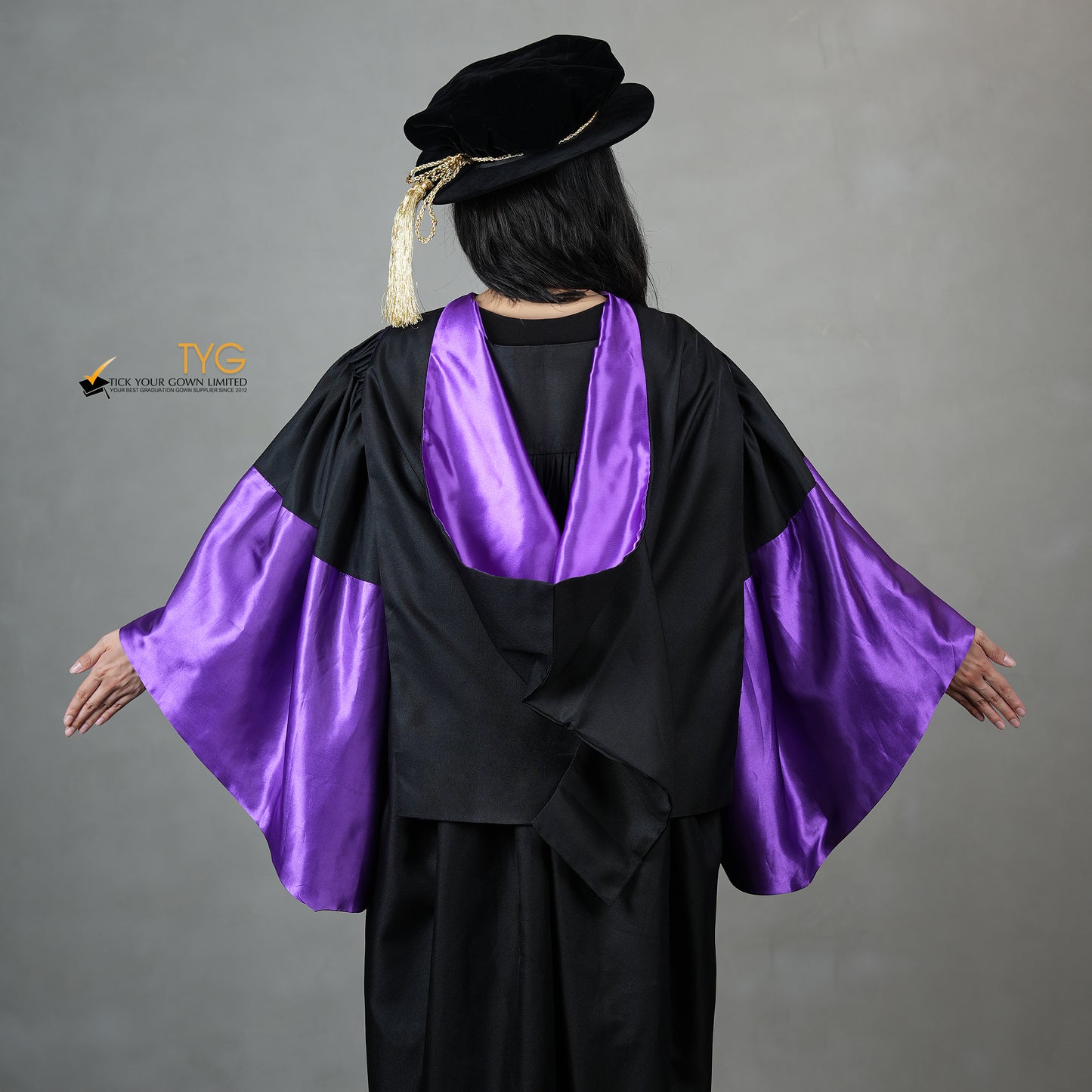 A detailed back view of a person wearing a graduation gown in black with purple satin panels, matching purple satin sleeve cuffs, and a black doctoral bonnet with gold tassel.
